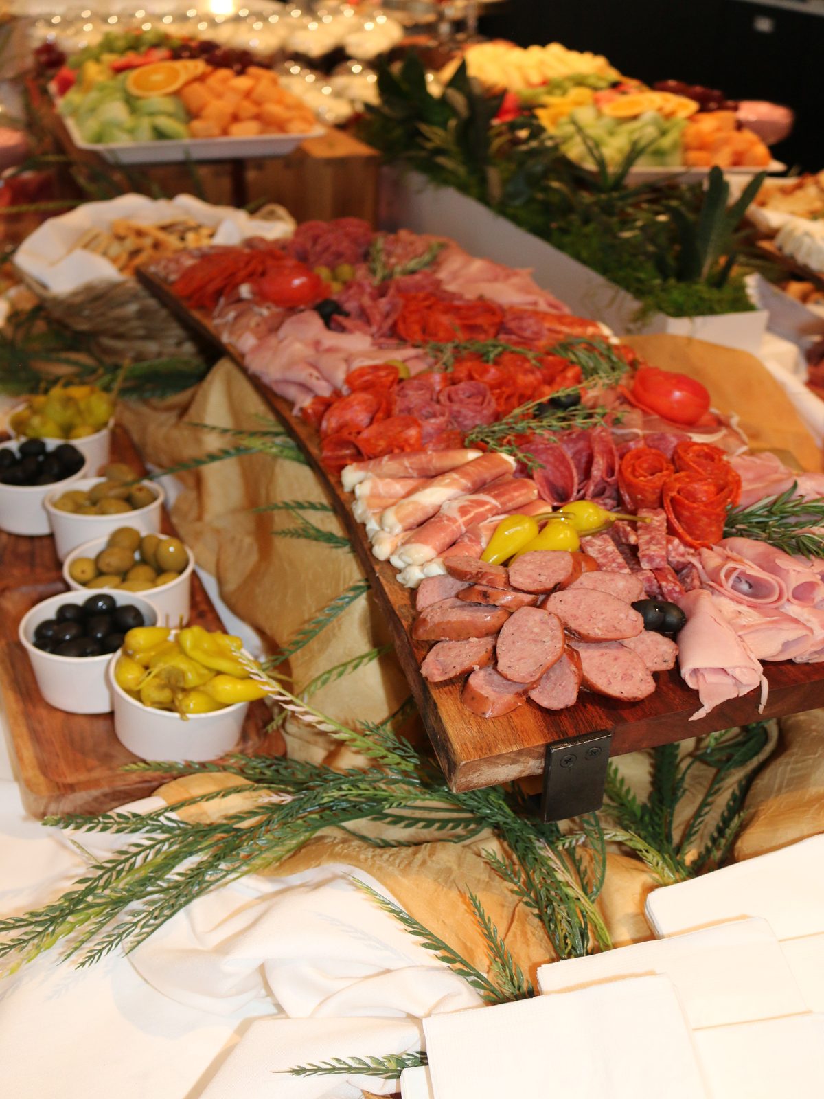Live-edge wooden board of rolled salami, prosciutto and sliced sausage with tomato roses and rosemary, alongside bowls of olives and pepperoncini
