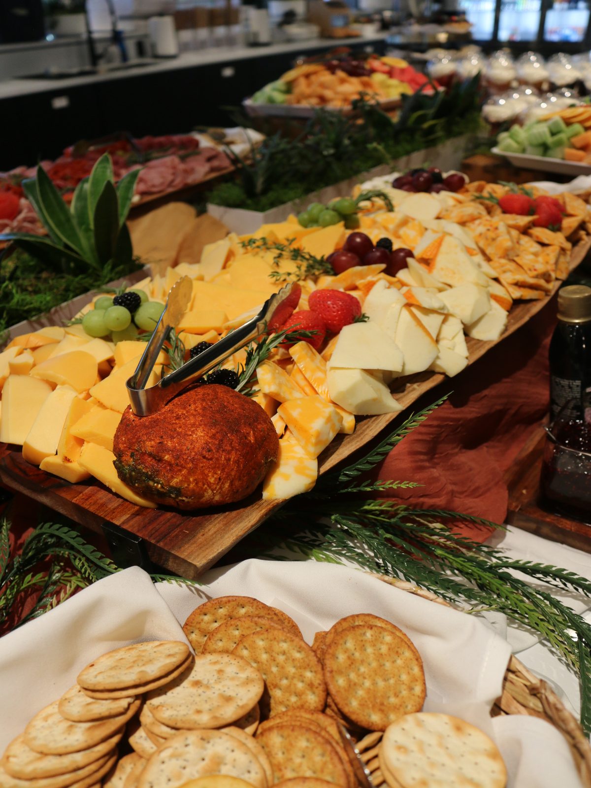 Wooden board of cubed and sliced cheddar and jack cheeses with a herb-crusted cheese ball, grapes and strawberries, beside a basket of crackers
