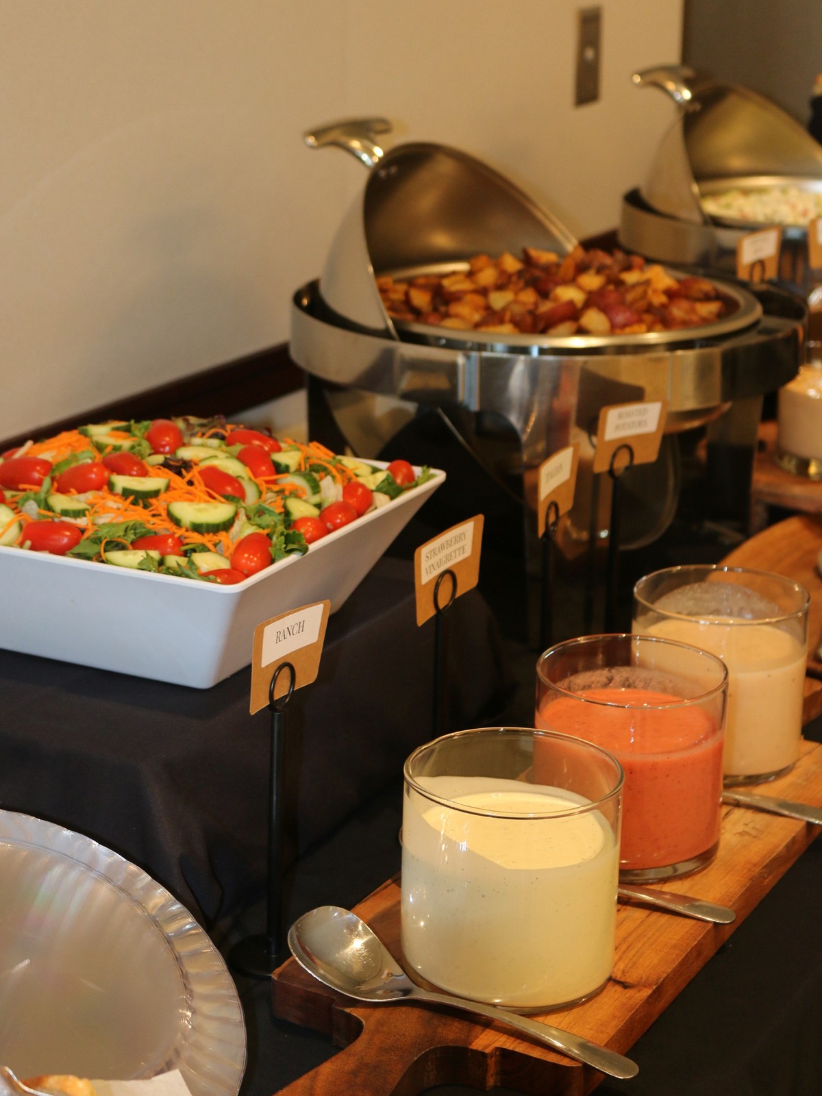 Buffet service line with a cucumber and cherry-tomato salad, a chafing dish of roasted potatoes and glasses of house dressings