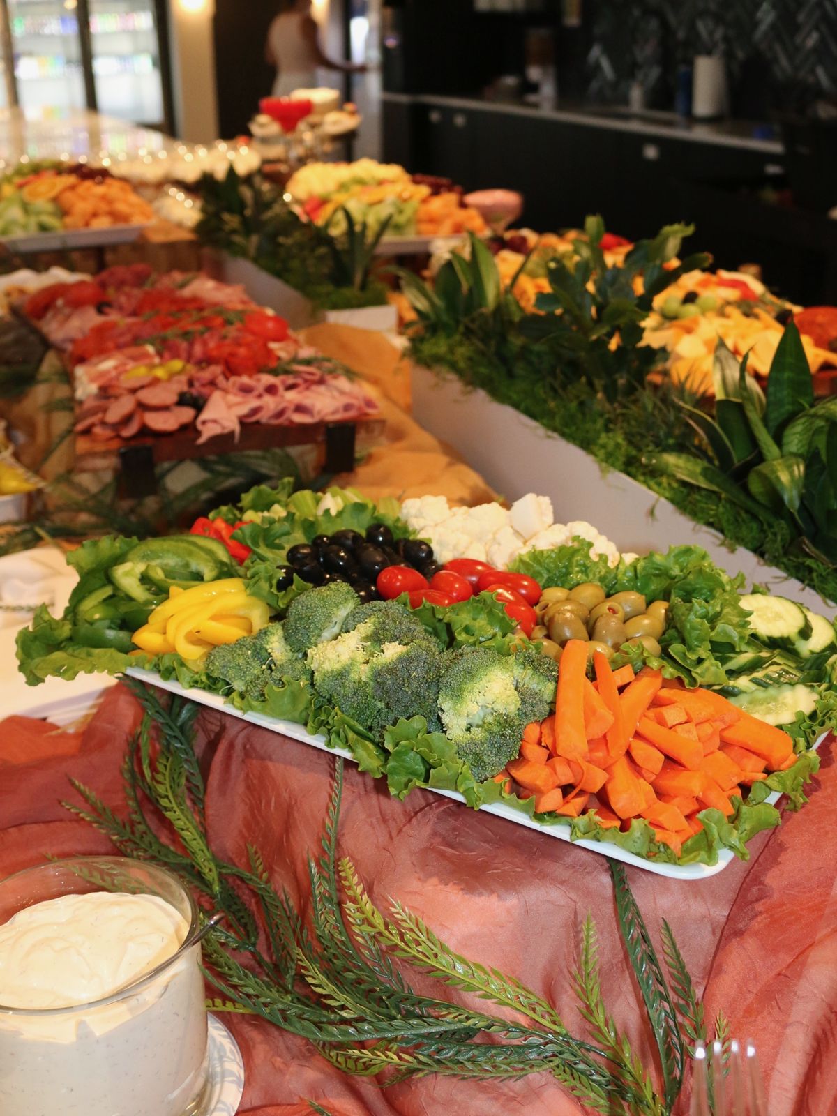 Vegetable tray of broccoli, carrots, peppers, olives and cauliflower on a bed of lettuce, beside a buffet line of sliced deli meats